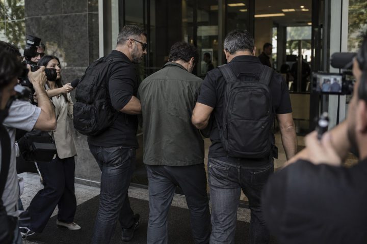Cena ilustrativa de uma pessoa conduzida por agentes, cercada por repórteres e câmeras.